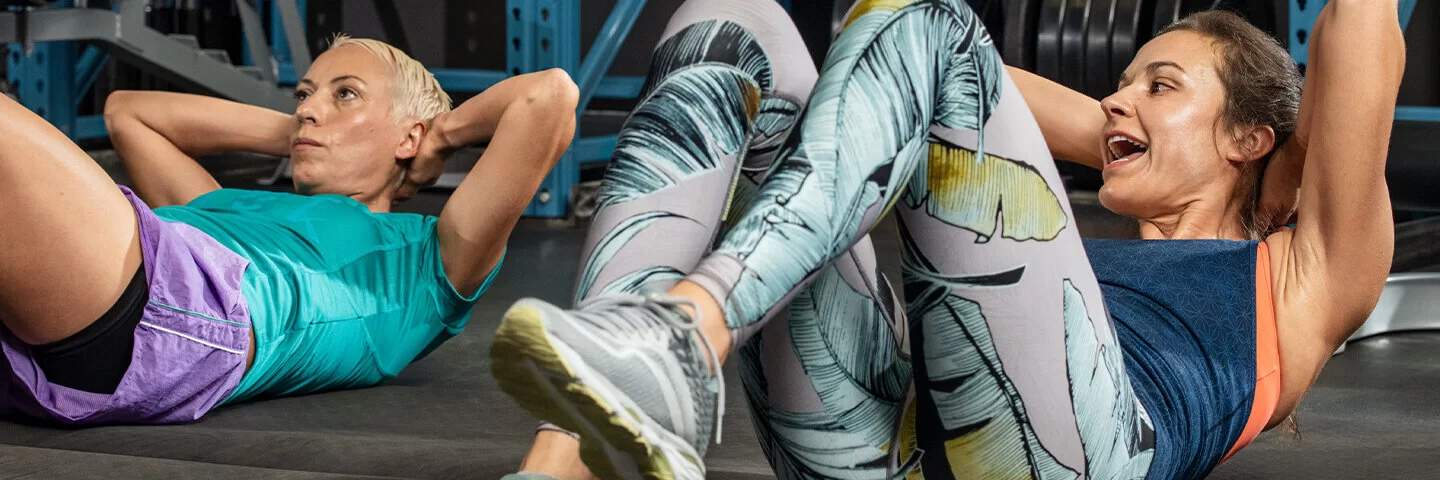 Das Bild zeigt zwei Frauen in Sportkleidung in einem Fitnessstudio, die Sit-ups machen.