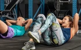 Das Bild zeigt zwei Frauen in Sportkleidung in einem Fitnessstudio, die Sit-ups machen.