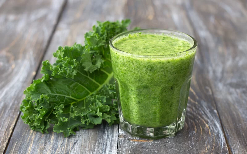 Auf einem Tisch steht ein Glas Grünkohlsmoothie und daneben liegt ein Grünkohlblatt.