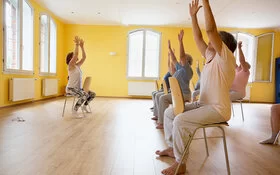 Eine Gruppe von Senioren und Seniorinnen macht unter Anleitung Gymnastik im Sitzen.