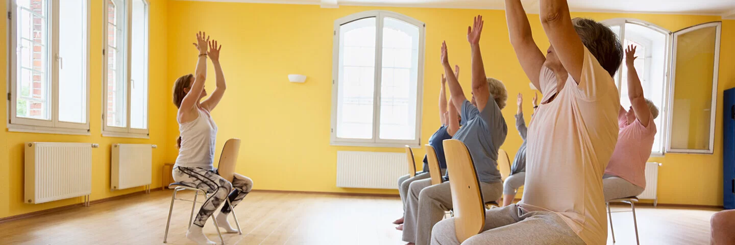 Eine Gruppe von Senioren und Seniorinnen macht unter Anleitung Gymnastik im Sitzen.