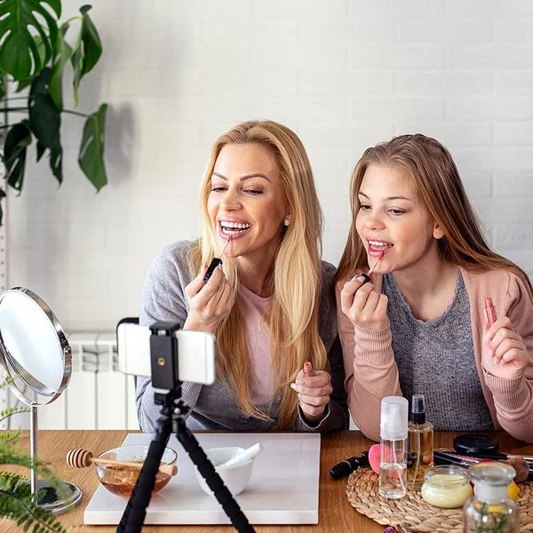 Eine Frau und ein Teenagermädchen sitzen an einem Tisch und schminken sich die Lippen. Auf dem Tisch stehen ein Schminkspiegel sowie ein Stativ mit einem Smartphone. Beide blicken zum Smartphone.