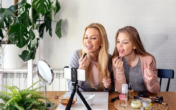 Eine Frau und ein Teenagermädchen sitzen an einem Tisch und schminken sich die Lippen. Auf dem Tisch stehen ein Schminkspiegel sowie ein Stativ mit einem Smartphone. Beide blicken zum Smartphone.