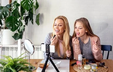 Eine Frau und ein Teenagermädchen sitzen an einem Tisch und schminken sich die Lippen. Auf dem Tisch stehen ein Schminkspiegel sowie ein Stativ mit einem Smartphone. Beide blicken zum Smartphone.