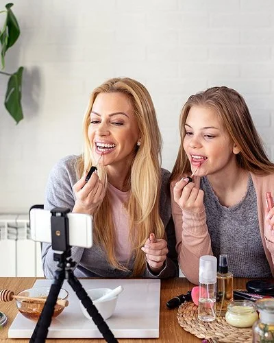 Eine Frau und ein Teenagermädchen sitzen an einem Tisch und schminken sich die Lippen. Auf dem Tisch stehen ein Schminkspiegel sowie ein Stativ mit einem Smartphone. Beide blicken zum Smartphone.