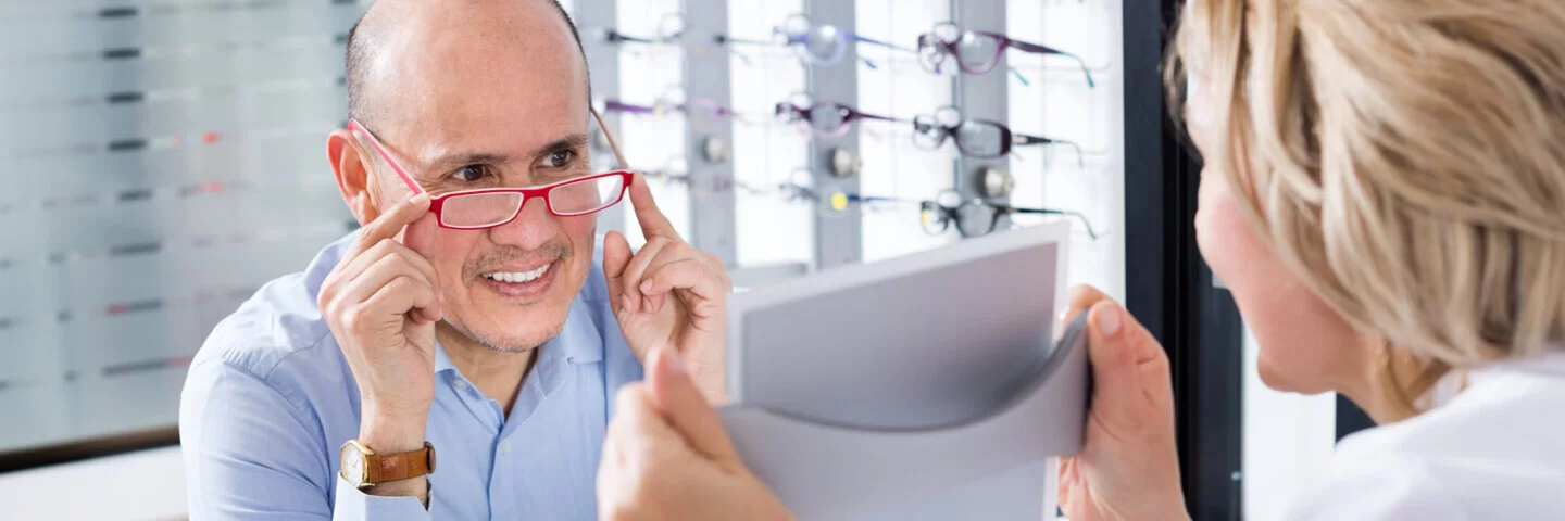 Ein Mann lässt sich von einer Optikerin eine neue Brille ziegen. Diese zählt zu den wichtigen Hilfsmitteln für Versicherte.