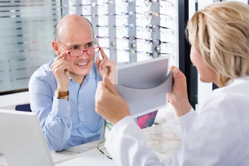 Ein Mann lässt sich von einer Optikerin eine neue Brille ziegen. Diese zählt zu den wichtigen Hilfsmitteln für Versicherte.