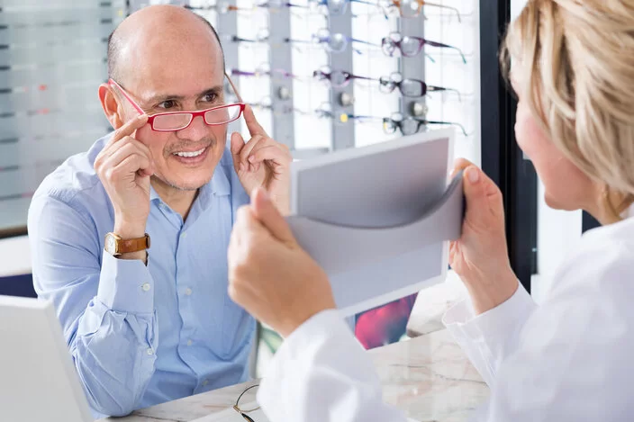 Ein Mann lässt sich von einer Optikerin eine neue Brille ziegen. Diese zählt zu den wichtigen Hilfsmitteln für Versicherte.