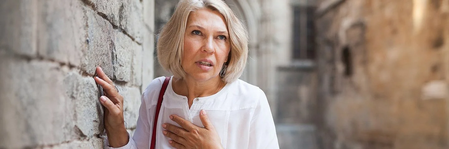 Eine Frau mit schulterlangen Haaren schaut angestrengt und hält sich auf der Straße mit einer Hand an die Brust, während sie sich mit der anderen an einer Steinmauer festhält.