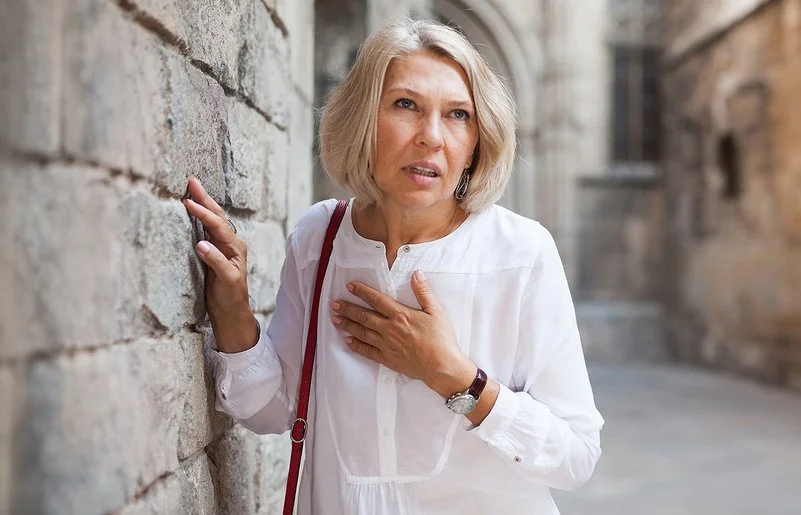 Eine Frau mit schulterlangen Haaren schaut angestrengt und hält sich auf der Straße mit einer Hand an die Brust, während sie sich mit der anderen an einer Steinmauer festhält.