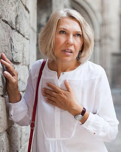 Eine Frau mit schulterlangen Haaren schaut angestrengt und hält sich auf der Straße mit einer Hand an die Brust, während sie sich mit der anderen an einer Steinmauer festhält.