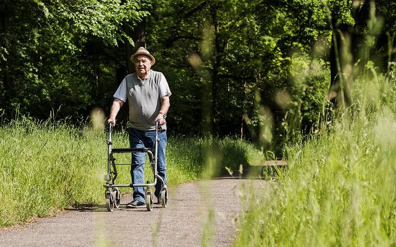 Ein älterer Mann spaziert mit einem Rollator auf einem Weg durch den Wald.