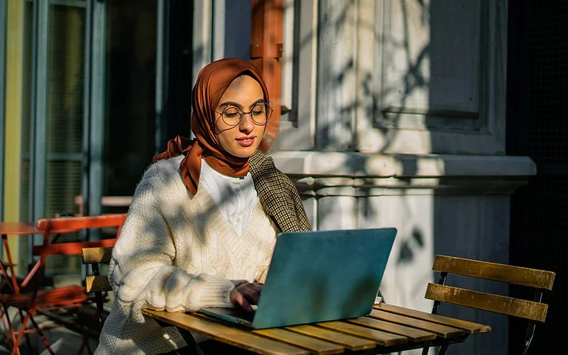 Eine junge Frau mit Kopftuch und Brille sitzt in einem Café am Laptop.