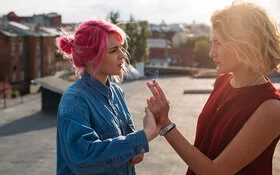 Zwei Teenagerinnen vor unscharfem Hintergrund: Eine hält eine Zigarette in der Hand, während die andere diese Hand von sich wegdrückt.