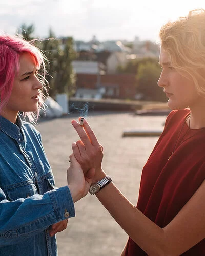 Zwei Teenagerinnen vor unscharfem Hintergrund: Eine hält eine Zigarette in der Hand, während die andere diese Hand von sich wegdrückt.