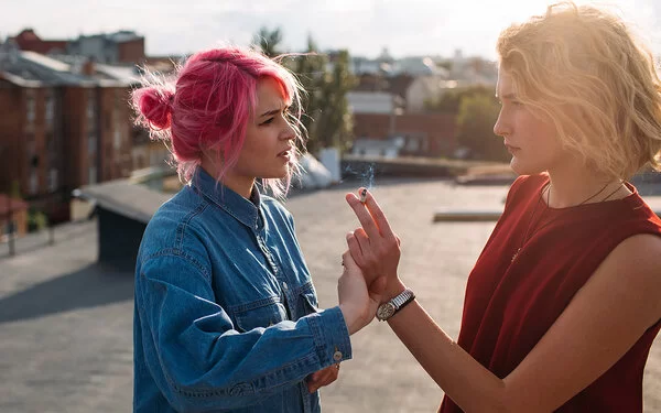 Zwei Teenagerinnen vor unscharfem Hintergrund: Eine hält eine Zigarette in der Hand, während die andere diese Hand von sich wegdrückt.