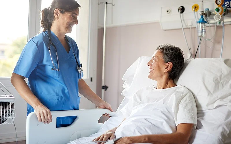 Eine Krankenschwester steht am Bett einer Patientin, die im Bett sitzt; beide sehen einander an und lächeln.