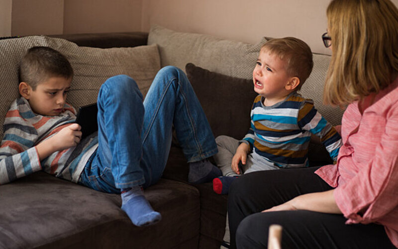 Ein kleiner Junge sitzt auf einem Sofa und weint, neben ihm eine ältere Frau, die ihn anschaut – gegenüber einem weiteren Jungen, der auf ein Smartphone blickt.