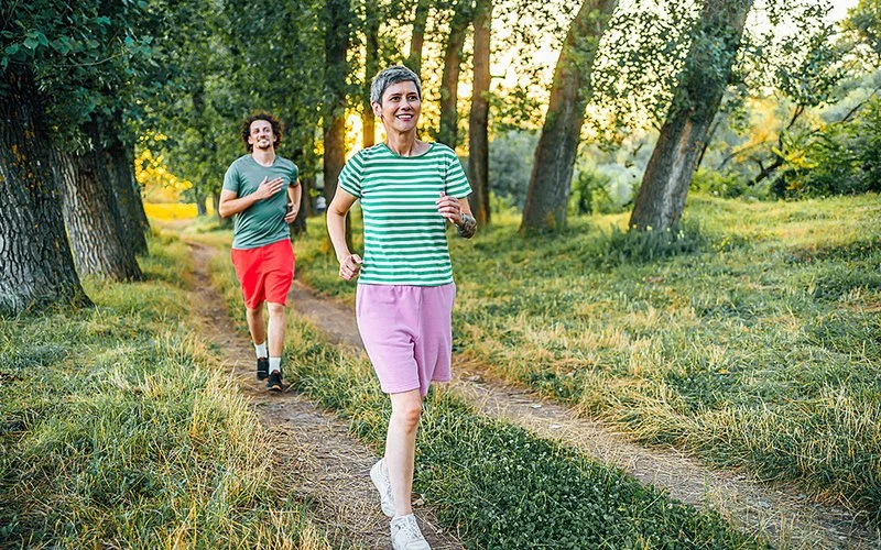 Eine schlanke, reife Frau in Sportkleidung läuft – gefolgt von einem männlichen Mitläufer – auf einem Weg im Freien, umgeben von Natur.