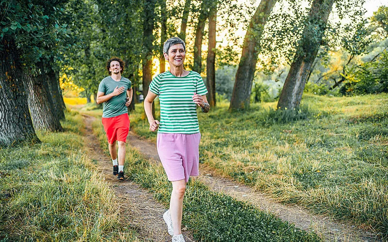 Eine schlanke, reife Frau in Sportkleidung läuft – gefolgt von einem männlichen Mitläufer – auf einem Weg im Freien, umgeben von Natur.