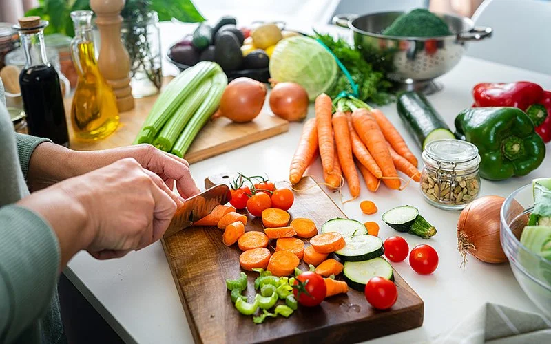 Die Hände einer Frau, die in der Küche vor der Arbeitsfläche steht. Auf der Arbeitsfläche liegen ein Schneidbrett sowie diverse Gemüse, darunter Karotten, Sellerie, Paprika, Zwiebeln und Tomaten. Im Hintergrund befinden sich ein Glas mit Kräutern, eine Flasche mit Essig und Öl, eine Pfeffermühle und weitere Küchenutensilien. Die Frau schneidet eine Karotte.