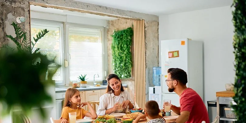 Eine fröhliche junge Familie sitzt an einem Esstisch in einer modern eingerichteten Küche. Im Bild zu sehen sind die Mutter und die Tochter, ihnen gegenüber sitzen Vater und Sohn. Auf dem Tisch stehen Brot und weitere Lebensmittel für ein gesundes Frühstück.