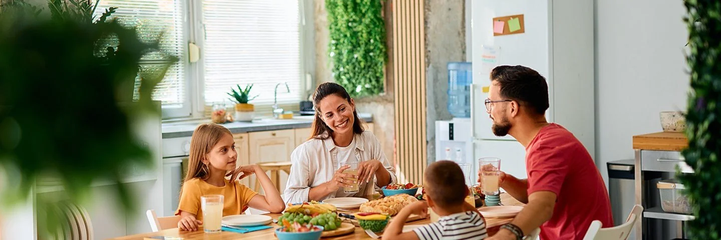 Eine fröhliche junge Familie sitzt an einem Esstisch in einer modern eingerichteten Küche. Im Bild zu sehen sind die Mutter und die Tochter, ihnen gegenüber sitzen Vater und Sohn. Auf dem Tisch stehen Brot und weitere Lebensmittel für ein gesundes Frühstück.