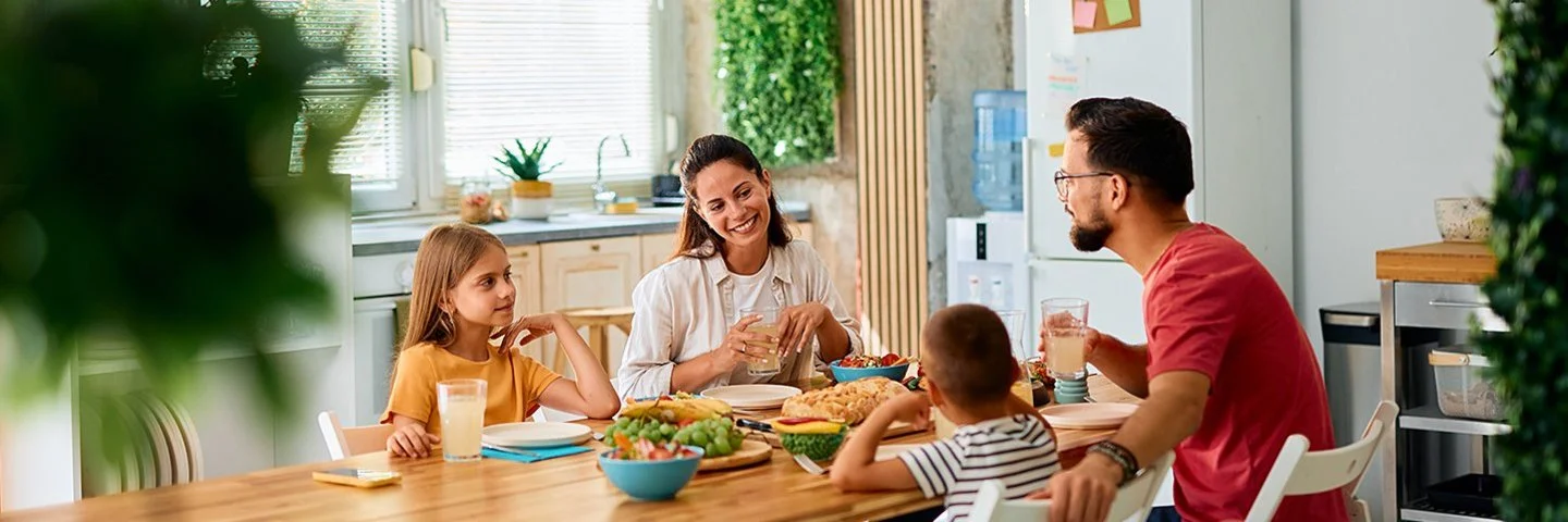 Eine fröhliche junge Familie sitzt an einem Esstisch in einer modern eingerichteten Küche. Im Bild zu sehen sind die Mutter und die Tochter, ihnen gegenüber sitzen Vater und Sohn. Auf dem Tisch stehen Brot und weitere Lebensmittel für ein gesundes Frühstück.