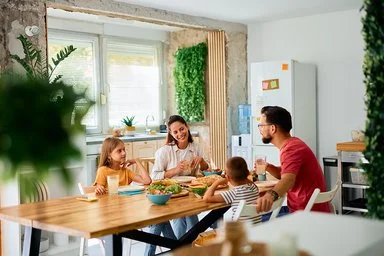 Eine fröhliche junge Familie sitzt an einem Esstisch in einer modern eingerichteten Küche. Im Bild zu sehen sind die Mutter und die Tochter, ihnen gegenüber sitzen Vater und Sohn. Auf dem Tisch stehen Brot und weitere Lebensmittel für ein gesundes Frühstück.