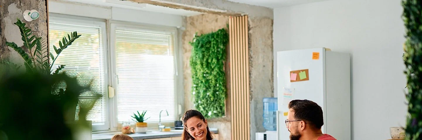 Eine fröhliche junge Familie sitzt an einem Esstisch in einer modern eingerichteten Küche. Im Bild zu sehen sind die Mutter und die Tochter, ihnen gegenüber sitzen Vater und Sohn. Auf dem Tisch stehen Brot und weitere Lebensmittel für ein gesundes Frühstück.