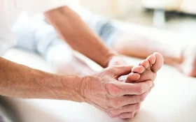 Eine Person liegt auf einer Untersuchungsliege, während ein älterer Arzt deren rechten Fuß in Händen hält und begutachtet.