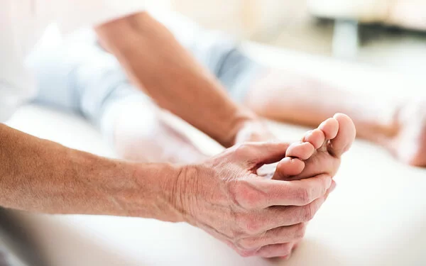 Eine Person liegt auf einer Untersuchungsliege, während ein älterer Arzt deren rechten Fuß in Händen hält und begutachtet.