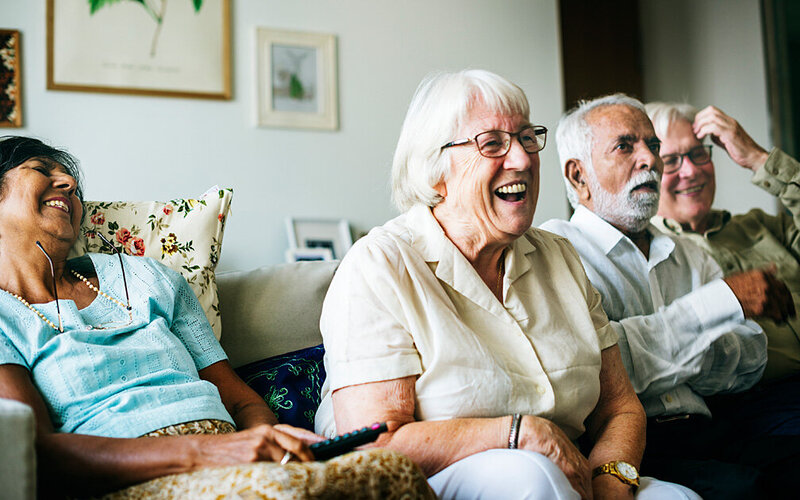 Vier ältere Menschen sitzen gemeinsam vergnügt auf dem Sofa – sie leben in einem betreuten Wohn-System.