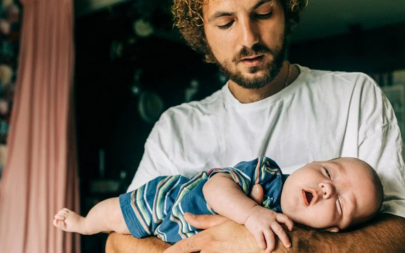 Mann hält ein Baby im Arm und blickt aufmerksam auf das Kind; das Baby liegt ruhig auf seinem Unterarm.