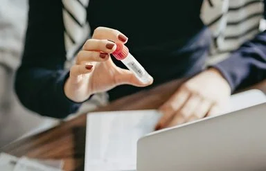 Eine junge Frau sitzt an Ihrem Computer und hält ein Proberöhrchen in die Kamera.