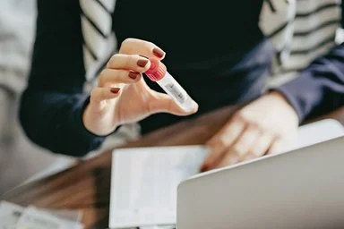Eine junge Frau sitzt an Ihrem Computer und hält ein Proberöhrchen in die Kamera.