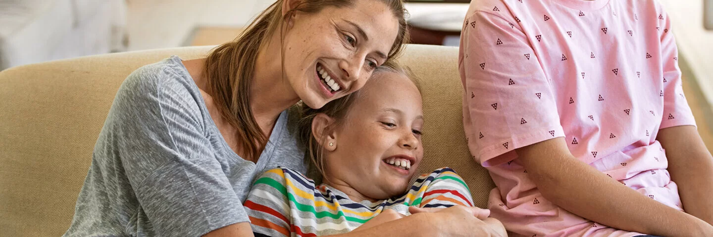 Eine Mutter sitzt mit ihren beiden Kindern lachend auf der Couch.