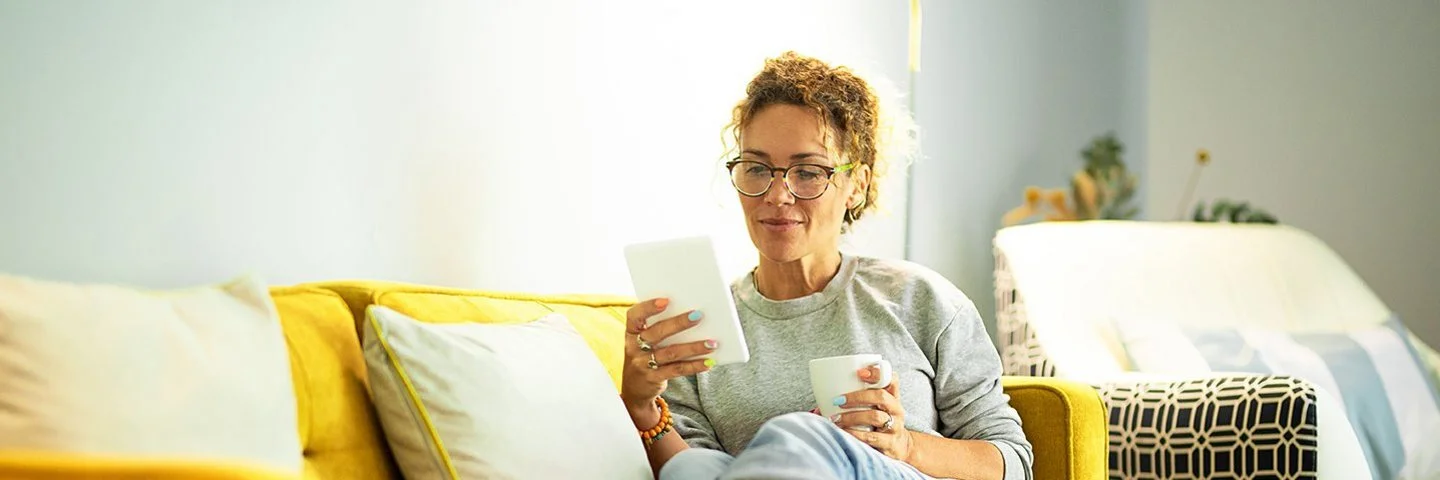 Frau mit Brille sitzt entspannt auf gelbem Sofa und liest von einem Tablet, sie hält eine Tasse in der anderen Hand.