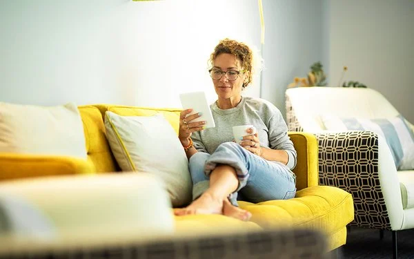Frau mit Brille sitzt entspannt auf gelbem Sofa und liest von einem Tablet, sie hält eine Tasse in der anderen Hand.