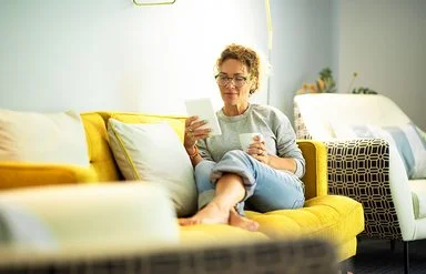 Frau mit Brille sitzt entspannt auf gelbem Sofa und liest von einem Tablet, sie hält eine Tasse in der anderen Hand.