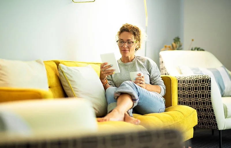 Frau mit Brille sitzt entspannt auf gelbem Sofa und liest von einem Tablet, sie hält eine Tasse in der anderen Hand.