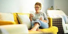 Frau mit Brille sitzt entspannt auf gelbem Sofa und liest von einem Tablet, sie hält eine Tasse in der anderen Hand.