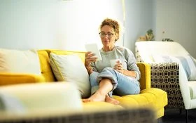 Frau mit Brille sitzt entspannt auf gelbem Sofa und liest von einem Tablet, sie hält eine Tasse in der anderen Hand.