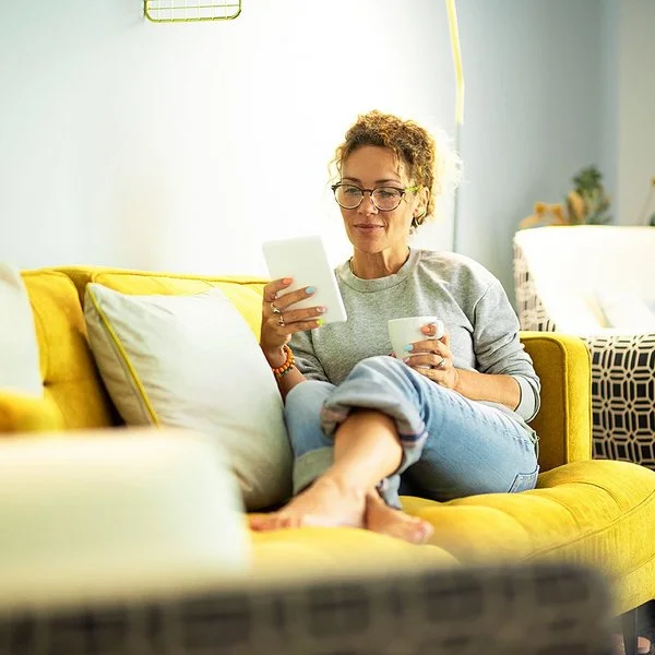 Frau mit Brille sitzt entspannt auf gelbem Sofa und liest von einem Tablet, sie hält eine Tasse in der anderen Hand.