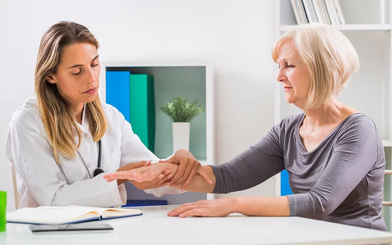 Eine junge Ärztin untersucht die rechte Hand einer älteren Patientin auf ein Ganglion.