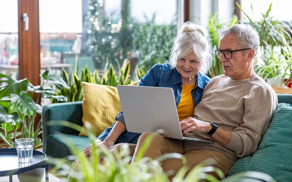 Ein Seniorenpaar sitzt mit einem Laptop auf der Wohnzimmercouch.