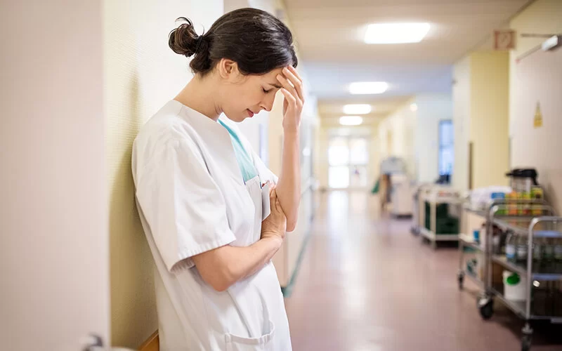 Eine erschöpfte junge Pflegerin lehnt sich im Gang eines Krankenhauses an die Wand und fasst sich an die Stirn.