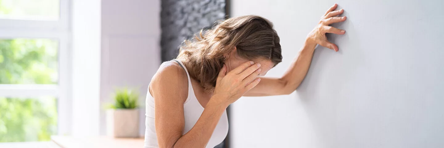 Eine junge Frau stützt sich mit links an der Wand ab, während sie sich mit rechts an die Schläfe fasst.