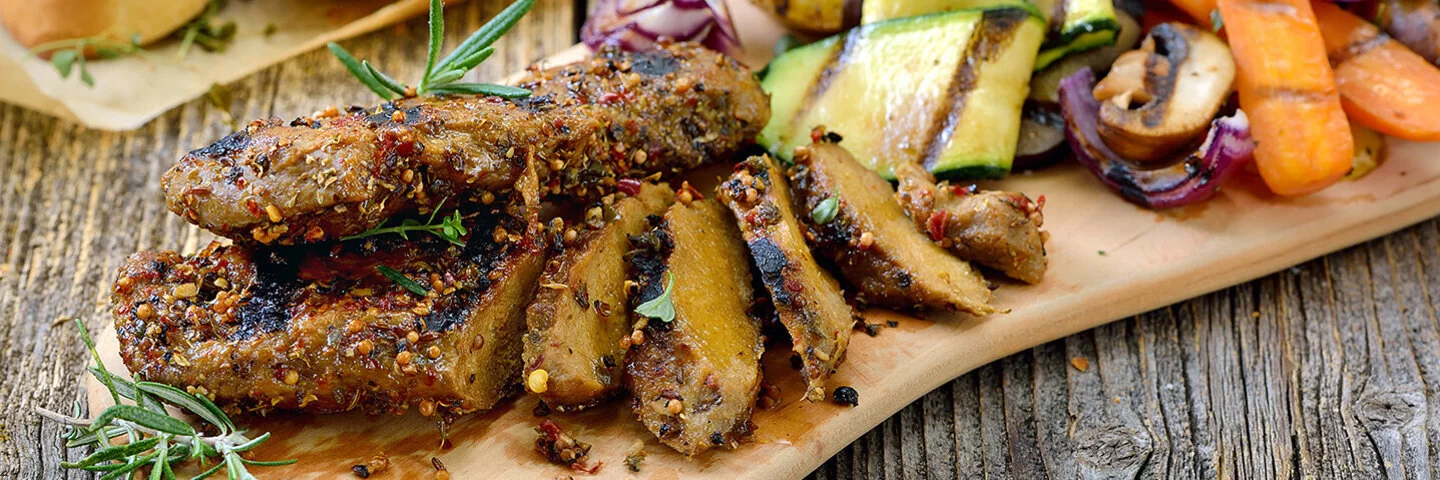 Auf einem Holzbrett liegen in Scheiben geschnittene Seitan-Steaks mit gegrillten Zucchini, Möhren, Zwiebeln und Maiskolben. Im Hintergrund sind gegrillte Baguettescheiben zu sehen.