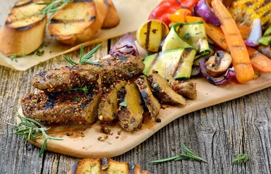 Auf einem Holzbrett liegen in Scheiben geschnittene Seitan-Steaks mit gegrillten Zucchini, Möhren, Zwiebeln und Maiskolben. Im Hintergrund sind gegrillte Baguettescheiben zu sehen.